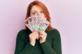 Beautiful redhead woman holding 100 new zealand dollars banknote in shock face, looking skeptical and sarcastic, surprised with Royalty Free Stock Photo