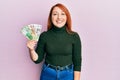 Beautiful redhead woman holding russian ruble banknotes looking positive and happy standing and smiling with a confident smile