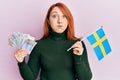 Beautiful redhead woman holding 20 swedish krona banknotes and sweden flag puffing cheeks with funny face Royalty Free Stock Photo