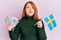 Beautiful redhead woman holding 20 swedish krona banknotes and sweden flag relaxed with serious expression on face Royalty Free Stock Photo