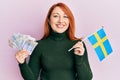 Beautiful redhead woman holding 20 swedish krona banknotes and sweden flag smiling with a happy and cool smile on face Royalty Free Stock Photo