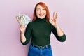 Beautiful redhead woman holding united kingdom pounds doing ok sign with fingers, smiling friendly gesturing excellent symbol