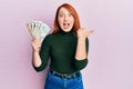 Beautiful redhead woman holding united kingdom pounds pointing thumb up to the side smiling happy with open mouth
