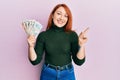 Beautiful redhead woman holding united kingdom pounds smiling happy pointing with hand and finger to the side