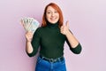 Beautiful redhead woman holding united kingdom pounds smiling happy and positive, thumb up doing excellent and approval sign