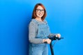 Beautiful redhead woman standing on modern scooter over blue background smiling with a happy and cool smile on face Royalty Free Stock Photo