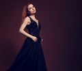 Beautiful redheaded young model with bright foxy hair posing in fashion chic black dress with long skirt on studio grey background Royalty Free Stock Photo