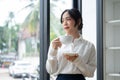 A beautiful Asian businesswoman sipping coffee while standing by the window in her office Royalty Free Stock Photo