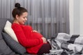 Beautiful relaxed pregnant woman sits at sofa, keeps hands on belly, enjoys comfort, being under plaid, has hair bun, poses in