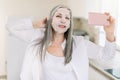 Beautiful retired grau haired lady in white shirt, standing at home kitchen and holding her cell phone, relaxing during