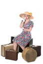 Beautiful retro blond wearing sun hat, seated with vintage suitcases, isolated on white