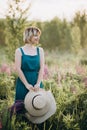 Beautiful romantic girl blonde in a dress in a field of purple flowers of lupins at dawn holds a hat in her hands Royalty Free Stock Photo