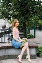 A beautiful romantic girl in a pink top and denim skirt holding a bouquet of roses and sitting on a bench in the city square Royalty Free Stock Photo