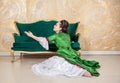 Beautiful sad woman in green rococo style medieval dress sitting on the floor near sofa and raises her hand Royalty Free Stock Photo