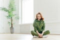 Beautiful satisfied slim woman with ginger hair, appealing appearance, sits crossed legs, wears green sweatsuit, keeps perfect