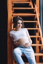 Beautiful saucy pregnant model with long brown hair in the white t-shirt and blue jeans sits on the wooden stairs and