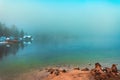 Beautiful scenic landscape of Lake Bohinj in foggy winter morning with old tree stump on shore and wooden boathouse with pier in