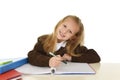 Beautiful schoolgirl in school uniform with blond hair smiling happy sitting on desk doing homework Royalty Free Stock Photo