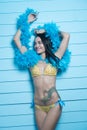 Beautiful seductive brunette woman wearing yellow bikini and blue feather boa smiling and looking into the camera over blue