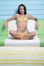 Beautiful seductive brunette woman wearing yellow bikini looking into the camera while sitting on the leather sofa over blue wall Royalty Free Stock Photo