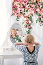 Beautiful senior woman with blonde hair looking at reflection in mirror in white studio with colorful flowers Royalty Free Stock Photo