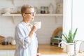 Beautiful senior woman with cup of coffee or rure water looking in window while enjoying new day at home Royalty Free Stock Photo