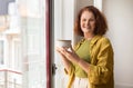 Beautiful senior woman enjoying cup of coffee while standing by the window Royalty Free Stock Photo