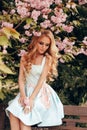 Beautiful sensual woman with blond hair in elegant clothes posing in garden with flowering sakura trees