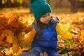 Beautiful serene baby girl in green woolen hat, yellow sweater and blue warm jacket sitting in fallen autumn dry leaves and