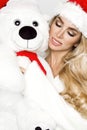 Beautiful blonde female model dressed in a Santa Claus hat embraces a white teddy bear in a red cap Christm