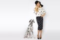 Beautiful, blonde woman in elegant polka dots and a hat, standing on a white background next to a dalmatian dog Royalty Free Stock Photo