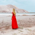 Beautiful cute girl with long blond hair in a long red evening dress standing in the desert near the mountains Royalty Free Stock Photo