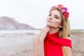 Beautiful cute girl with long blond hair in a long red evening dress with a wreath of roses and orchids in her hair standing Royalty Free Stock Photo