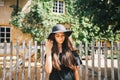 Beautiful girl brunette with brown eyes in a black dress and a black hat with big brims against the backdrop of a wooden fenc Royalty Free Stock Photo