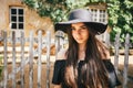 Beautiful girl brunette with brown eyes in a black dress and a black hat with big brims against the backdrop of a wooden fenc Royalty Free Stock Photo