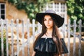 Beautiful girl brunette with brown eyes in a black dress and a black hat with big brims against the backdrop of a wooden fenc Royalty Free Stock Photo