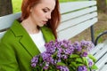 Beautiful girl with long hair in ryzhymi green coat sitting on a park bench with a bouquet of lilac in the hands of Royalty Free Stock Photo