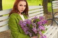 Beautiful girl with long hair in ryzhymi green coat sitting on a park bench with a bouquet of lilac in the hands of Royalty Free Stock Photo