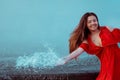 Beautiful sexy girl in a red dress with long hair laughs. Woman dancing near a fountain in the rain with wet hair