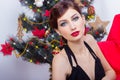 Beautiful happy smiling young woman in evening dress with bright makeup with red lipstick sitting near the Christmas tree Royalty Free Stock Photo