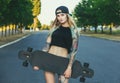 A beautiful, hipster blonde with blue hair in tattoo stands with a longboard Royalty Free Stock Photo