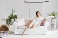 Beautiful and long legged blonde model girl in fashionable glasses and in stylish satin pajamas sits on the white Royalty Free Stock Photo