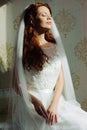 Beautiful redhair lady in elegant white wedding dress. Fashion portrait of model indoors. Beauty woman sitting near her bed Royalty Free Stock Photo