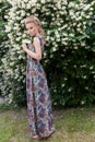 Beautiful tender blonde girl in a long dress with evening hairstyle standing in the garden near a flowering aromatic tree Royalty Free Stock Photo