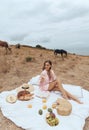 Beautiful sexy woman with blond hair in elegant dress and accessories having picnic with fantastic landscape and horses on