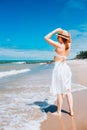 Beautiful sexy woman wearing straw hat, white swimsuit and skirt walking along the surf line on beach. Summer holidays in tropics Royalty Free Stock Photo