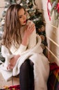 Beautiful sexy woman with Xmas tree in background sitting on elegant chair in cozy scenery. Portrait of girl posing Royalty Free Stock Photo