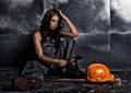 Beautiful tired miner worker sitting on a floor on steel background, pickaxe and orange helmet lies nearby