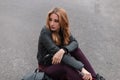 Beautiful sexy young woman in black leather jacket with a backpack in trendy pants relaxes sitting on the road in the city Royalty Free Stock Photo
