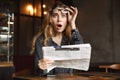 Beautiful shocked excited young woman sitting in cafe indoors reading newspaper